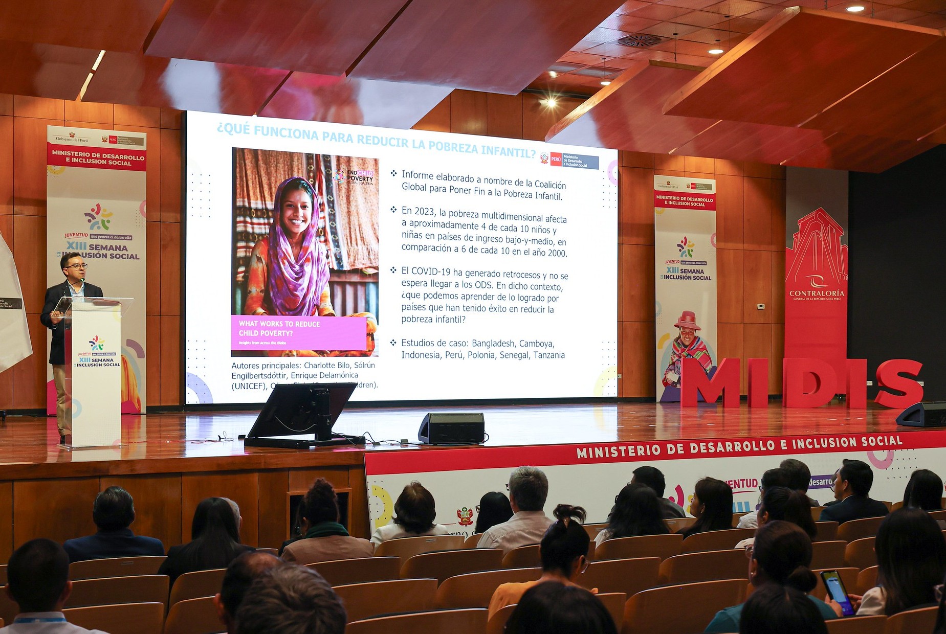 Alan Sanchez stood on a stage behind a lectern. To the right of him is a large screen showing a slide show and an image of a smiling young Indian woman.