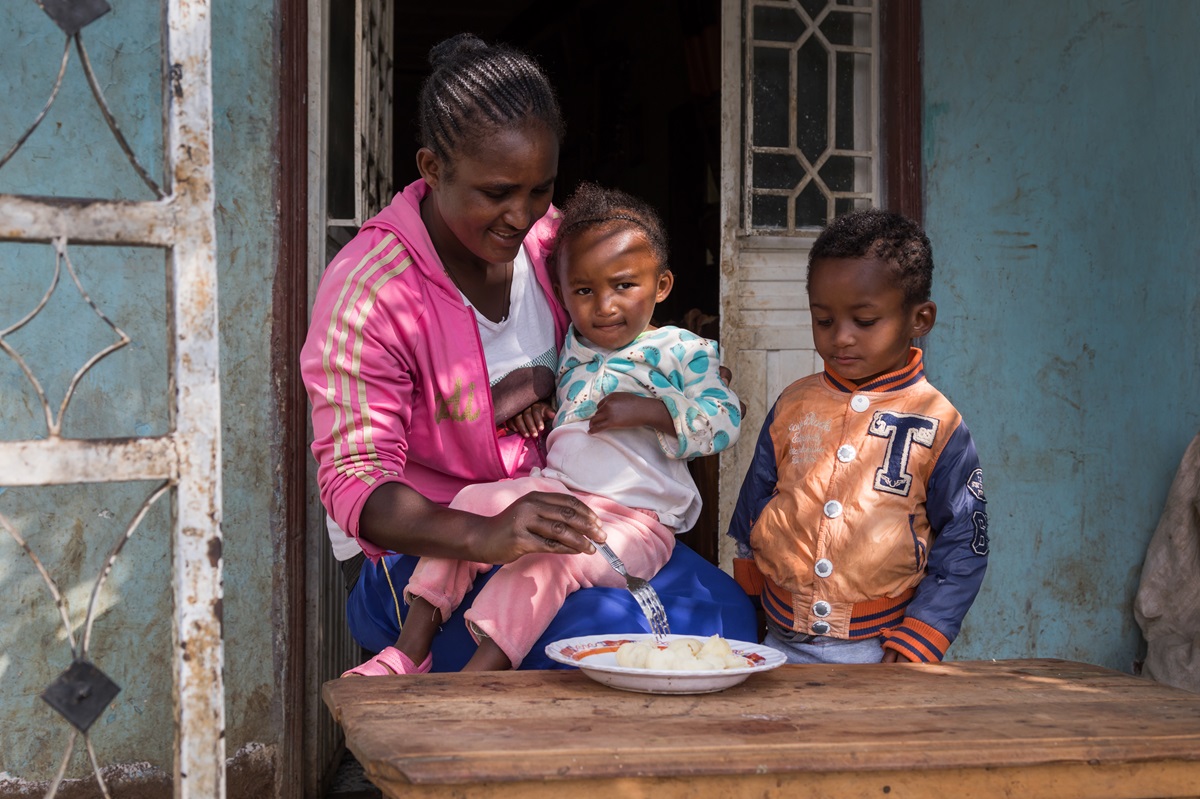 Ethiopian mother feeding her two young children whilst sat outside their home.