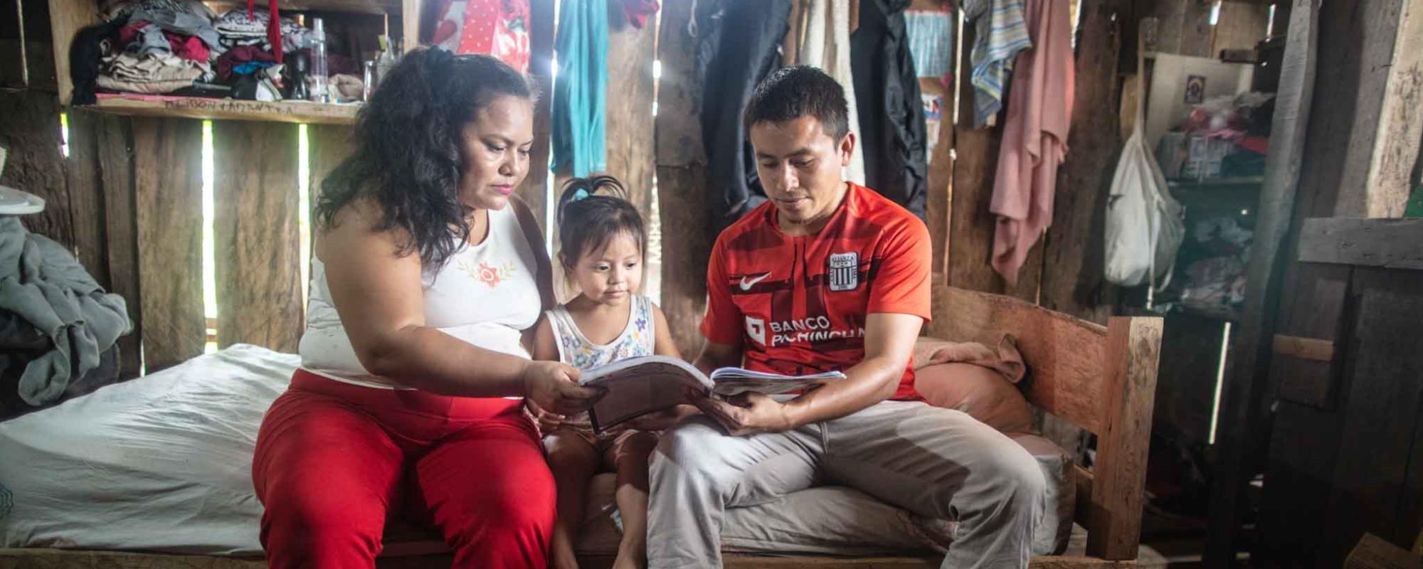 Mother and father sat on a wooden bed reading a book to their toddler daughter who is seated between them. Wall of the house behind them is rustic wooden panels through which you can see the light. There are clothes hanging from the wall.