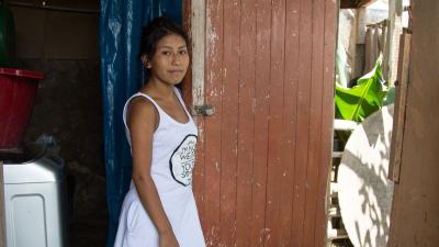 Young Peruvian woman standing inside her house holding the door open to her outside.