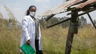 Women standing by a shack with a lab coat and face mask on 